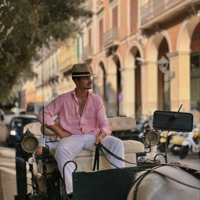 Adult male in a hat sits on a horse-drawn carriage on a European city street with old buildings and parked scooters on a sunny day.