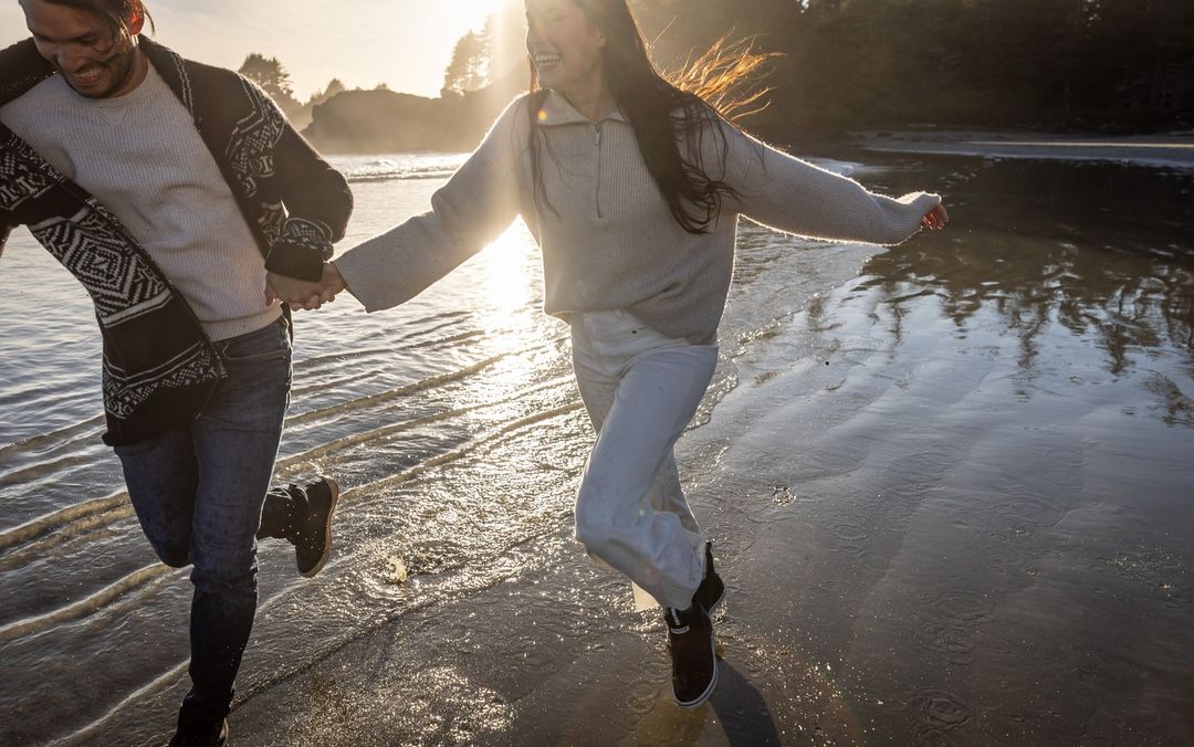 Couple running on a sandy beach at sunset. Woman in white sweater and jeans leads man in jeans and patterned sweater through the water.