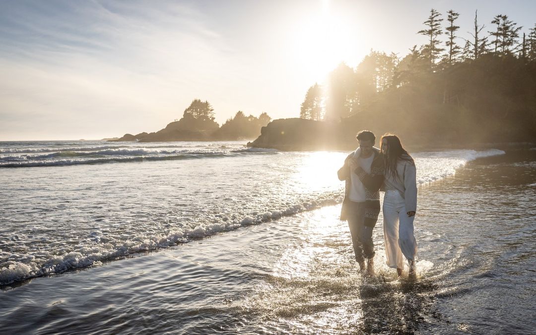 A couple strolls through surf at sunset on a sandy beach with gentle waves and a treed shoreline silhouetted in the background.