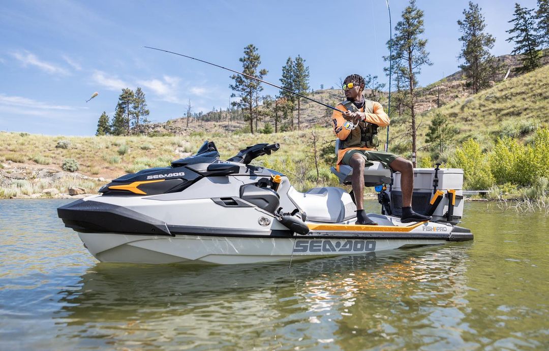 An adult male fishing from a Sea-Doo Fish Pro personal watercraft on a lake with a grassy, mountainous shoreline under a blue sky.