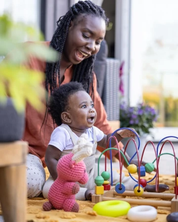Smiling African mother watches infant playing with wooden bead maze toy outdoors on mustard blanket with a pink crocheted stuffed animal.