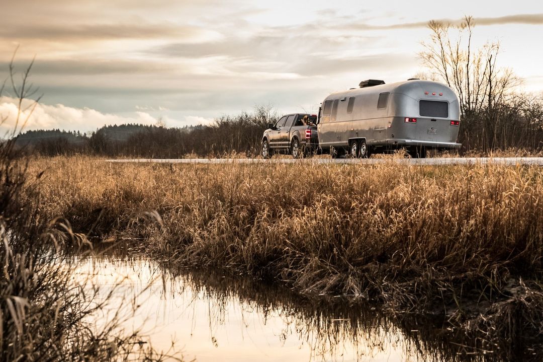Silver Airstream trailer towed by a pickup truck on a country road with tall grassy fields and moody skies at sunset.