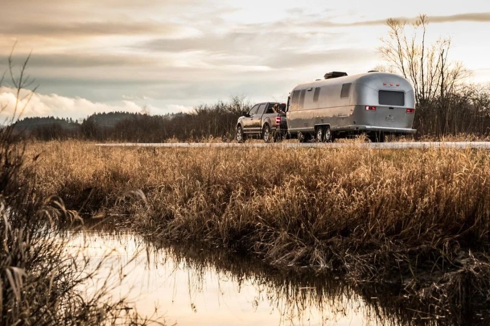 Silver Airstream trailer towed by a pickup truck on a country road with tall grassy fields and moody skies at sunset.