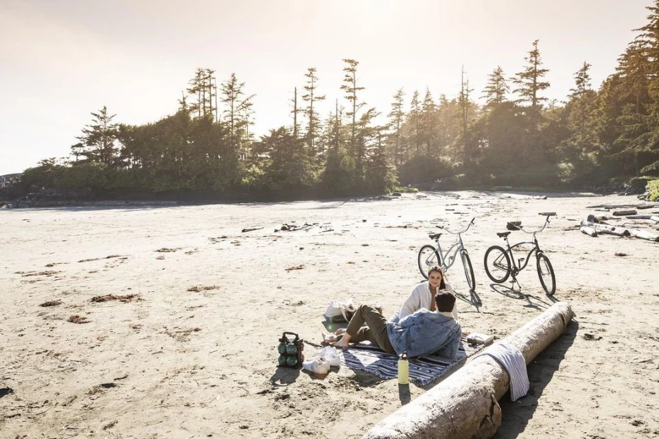 Couple relaxing on sandy beach with bicycles and trees in background under bright sky. Picnic scene with blanket, cooler and flask.