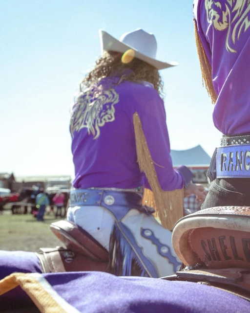 Cowboy riders in purple shirts and chaps at an outdoor event, showcasing traditional western attire and horsemanship under a clear sky.