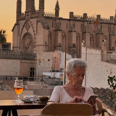 Elderly woman using a smartphone at a rooftop cafe, with a beer and snacks on the table, and a cathedral in the background.