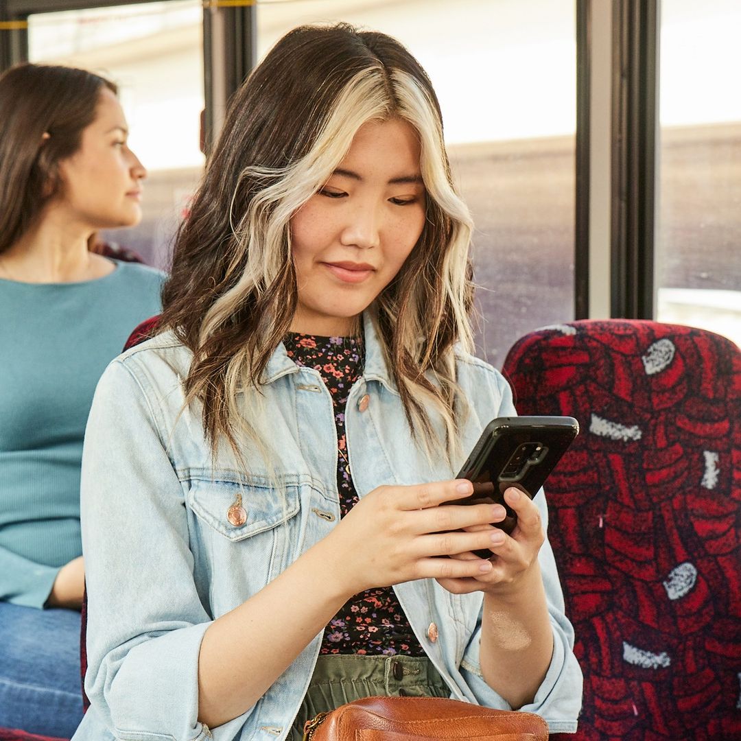 Young woman with blonde-streaked hair on a bus, looking at her smartphone, wearing a jean jacket and floral top, with another passenger visible.