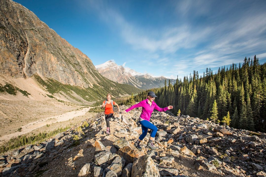 Two adult females hike up a rocky trail in the mountains under a blue sky with scattered clouds and evergreen trees.