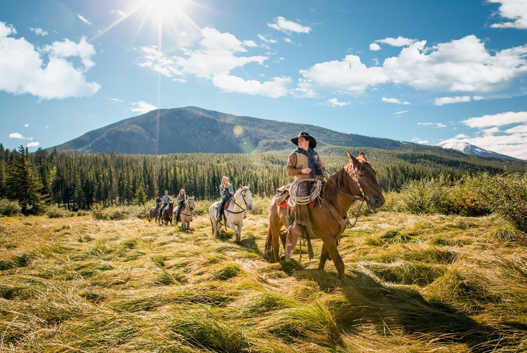 Group horseback riding tour in a grassy field beneath blue sky and mountain backdrop. Riders in hats enjoy sunny outdoor adventure.