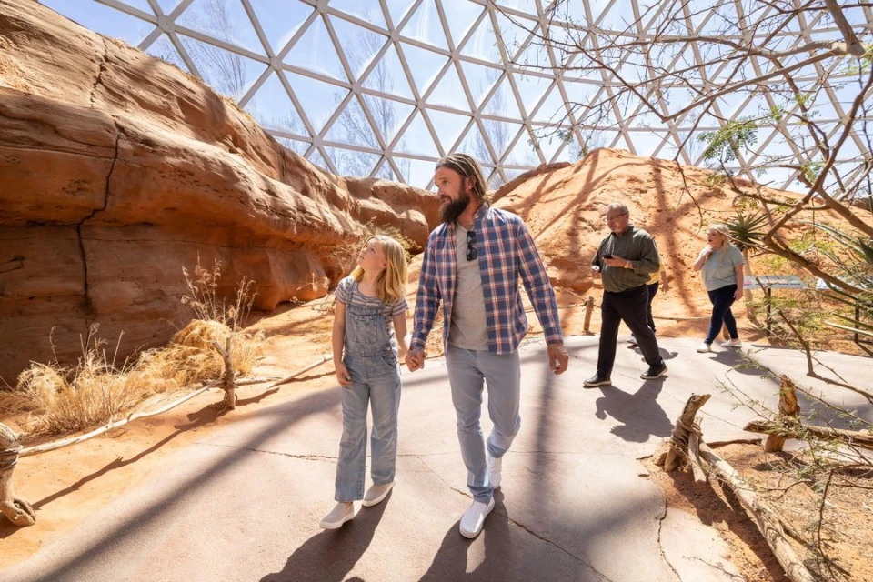A man and a child holding hands walking through an indoor desert exhibit with other visitors and desert plants under a geodesic dome.