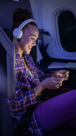 Woman smiles, using her phone, while seated on an airplane wearing headphones, a colorful plaid jacket, and violet pants.