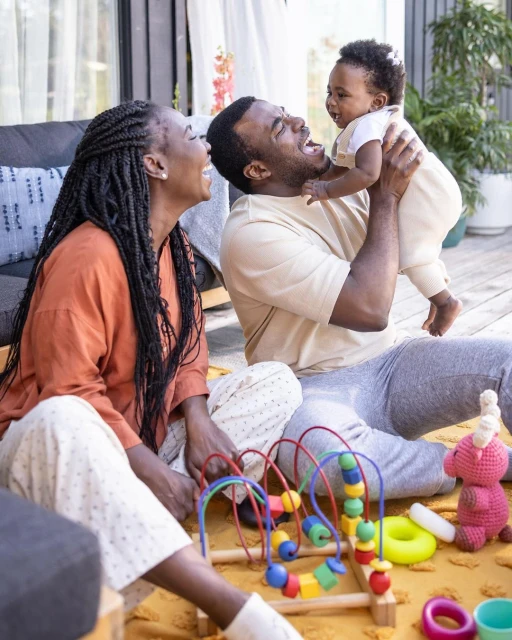 Happy African-American family of three smiling on a wooden deck with a baby held high in the air and toys on a yellow rug.