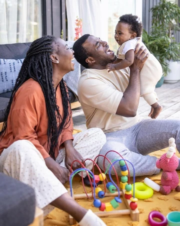 Happy African-American family of three smiling on a wooden deck with a baby held high in the air and toys on a yellow rug.