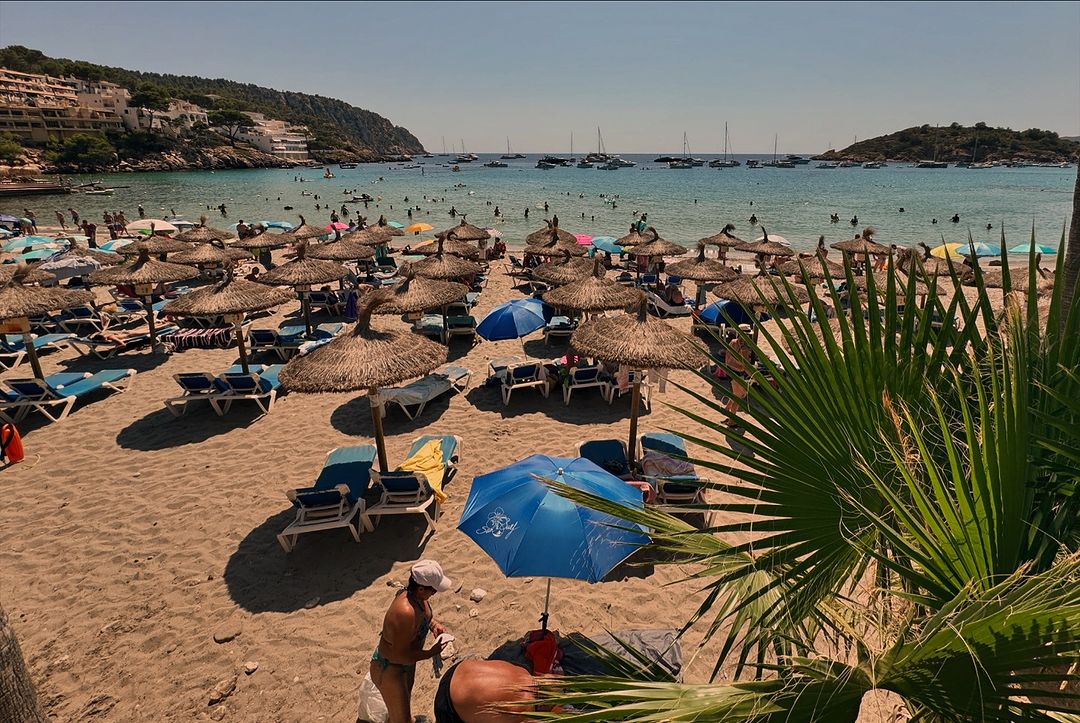 Sandy beach with straw umbrellas and lounge chairs facing turquoise water with boats, crowded with people relaxing on a sunny day.