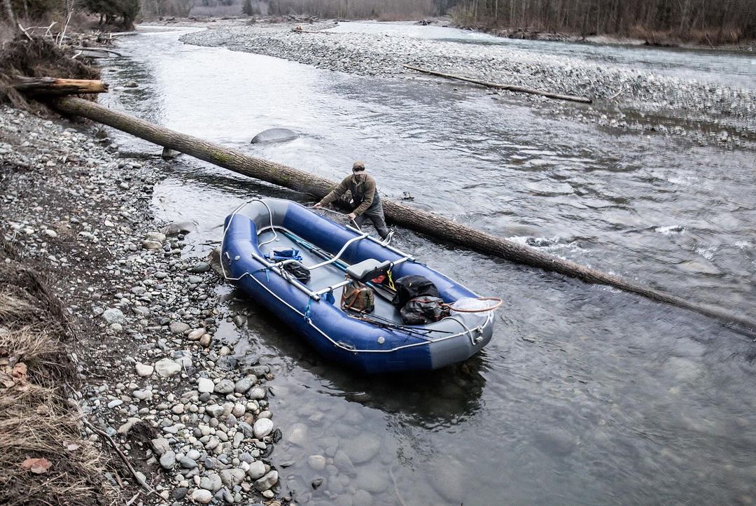 Man with blue inflatable raft and supplies in a rocky riverbed with a fallen log. Water flows around the raft and the shore is rocky.