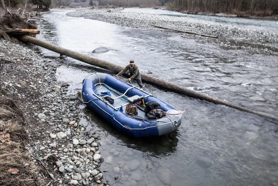 Man with blue inflatable raft and supplies in a rocky riverbed with a fallen log. Water flows around the raft and the shore is rocky.