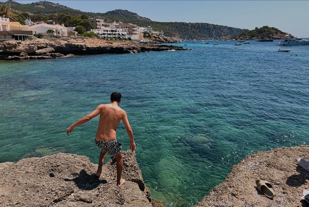 Young man standing on a rocky cliff overlooking turquoise water with boats, buildings, and mountains in the background on a sunny day.