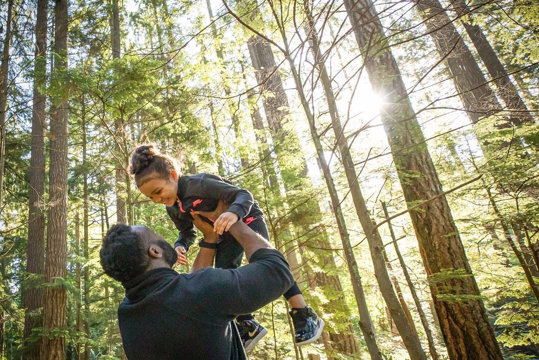 Father and daughter enjoying a sunny day in the forest, with the father holding the child up high among tall trees.