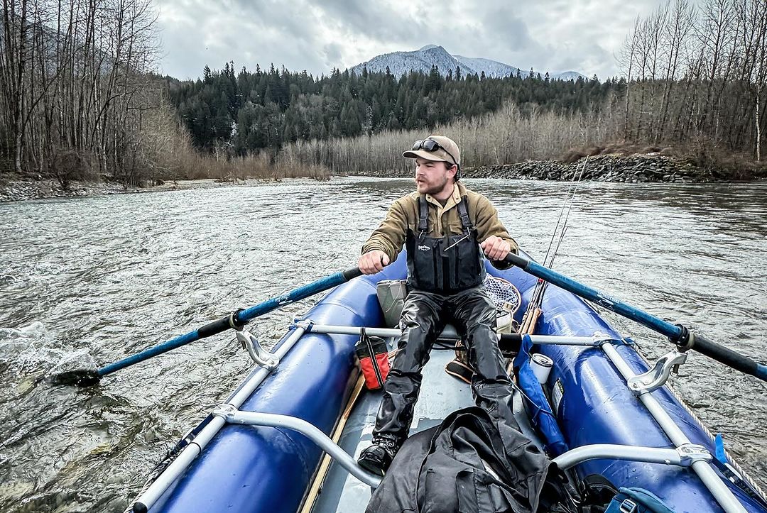 Man rows a blue inflatable raft on a river with mountain backdrop, wearing fishing gear, and captured at eye-level on a cloudy day.