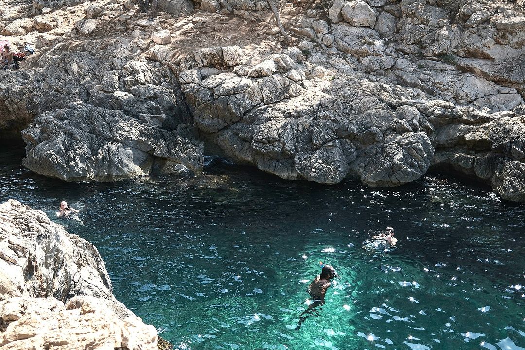 Overhead shot of people swimming in the teal water surrounded by light-grey cliffs and rock formations on a sunny day.