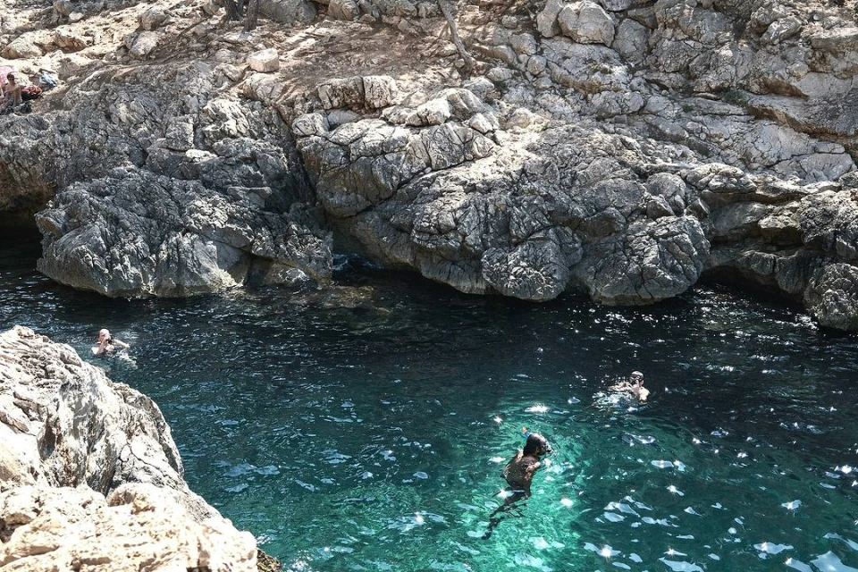 Overhead shot of people swimming in the teal water surrounded by light-grey cliffs and rock formations on a sunny day.