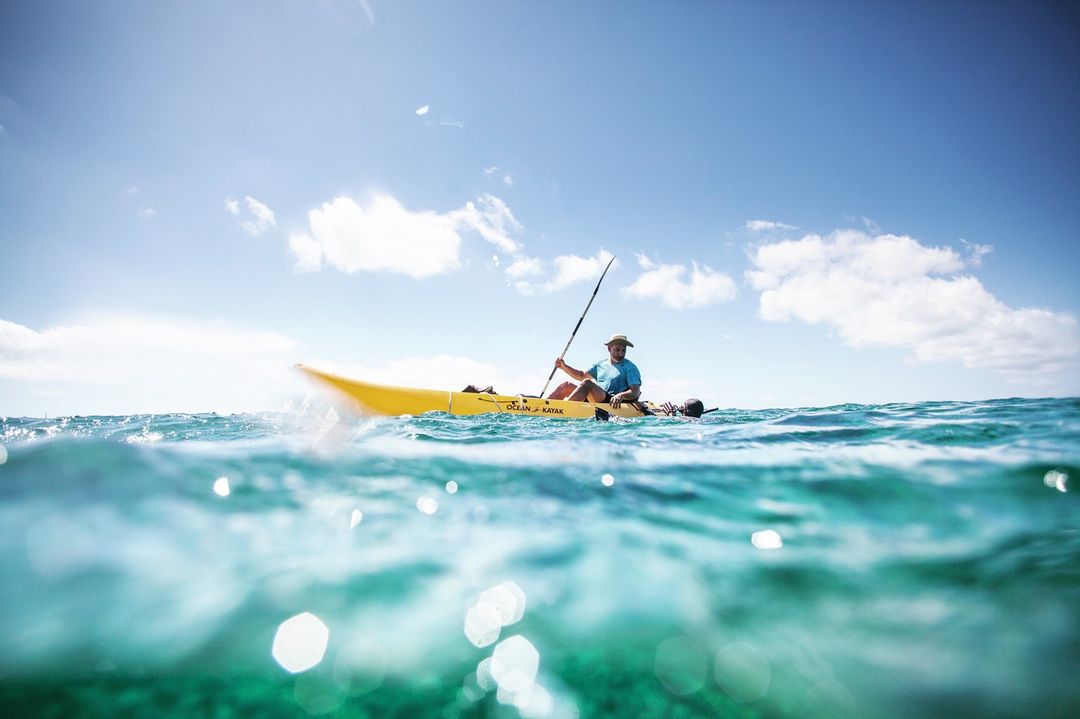 A man paddles a yellow Ocean Kayak on the ocean under a bright blue sky with light clouds in this sunny outdoor watersport image.
