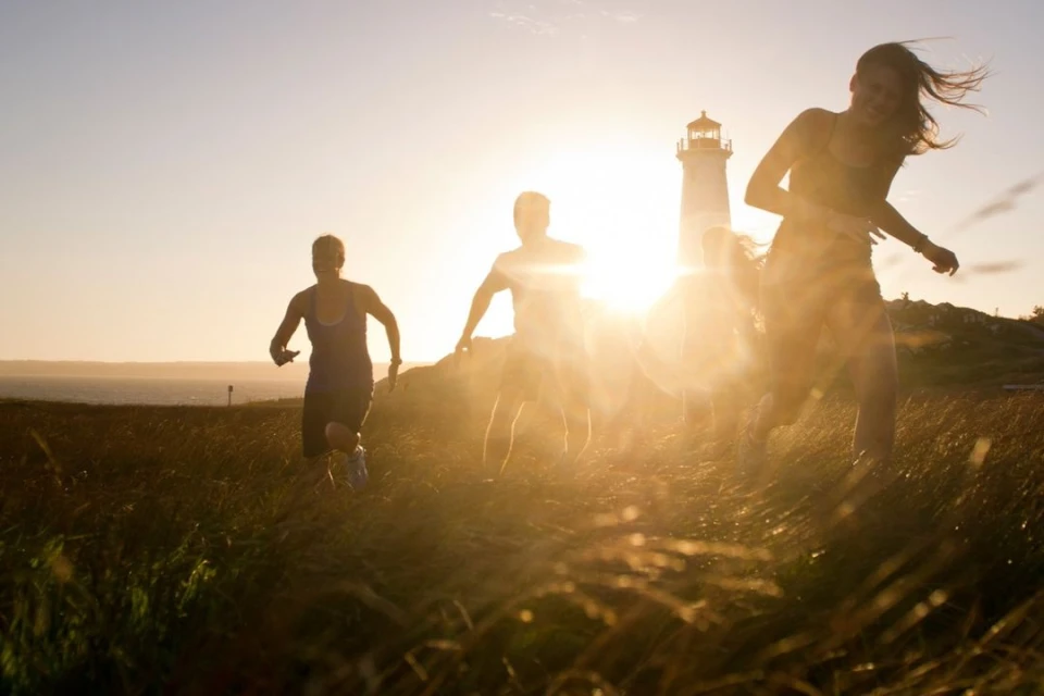 A group of people running toward the camera in a grassy field at sunset with a lighthouse visible in the background.
