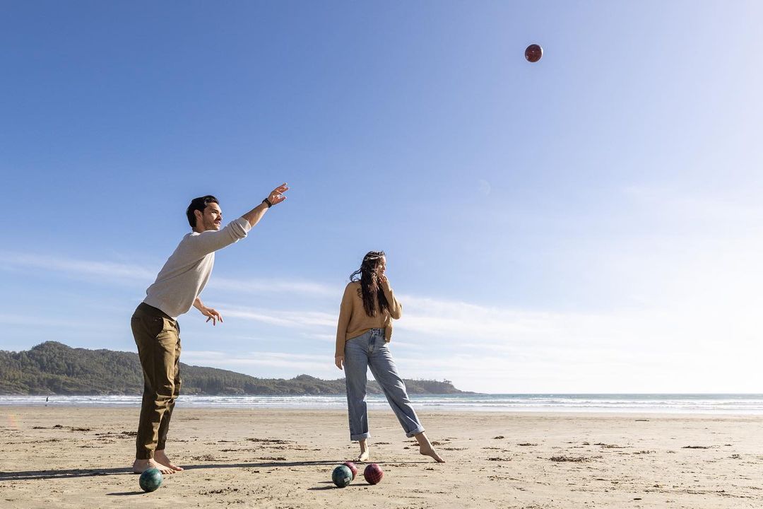 Couple playing bocce ball on sandy beach, man throwing ball, woman watching with blue sky, ocean, and trees in background during daytime.