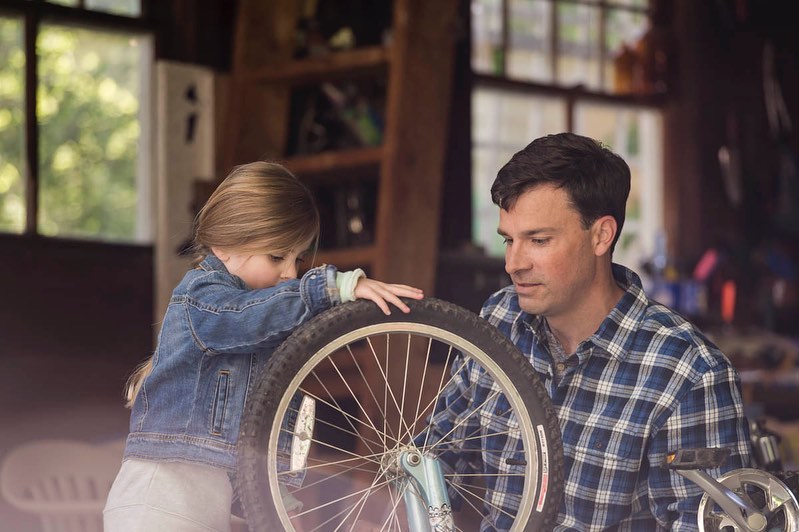 A dad and his daughter working on a bicycle wheel together inside a garage, with natural light coming in.