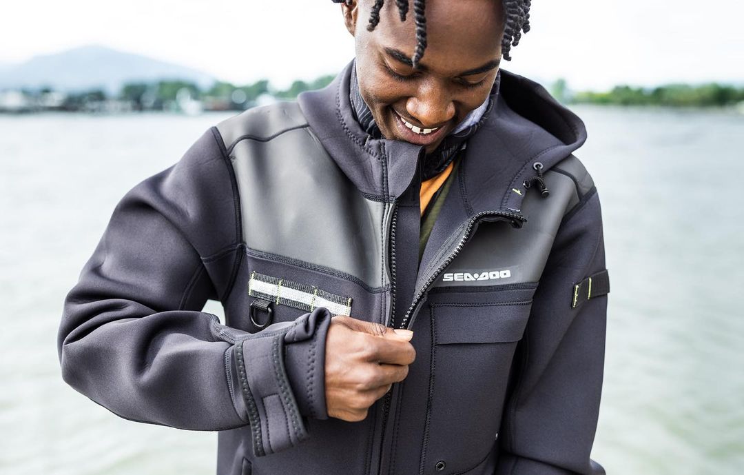 A smiling man with braided hair outdoors adjusts the zipper of his black and grey hooded jacket with the Seadoo logo visible.