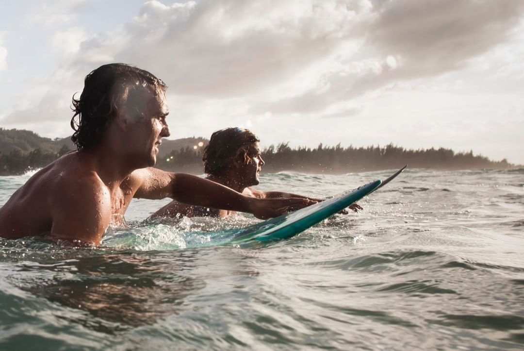Two adult male surfers paddle out on a turquoise surfboard in ocean waves near a forested coast under a bright, cloudy sky.