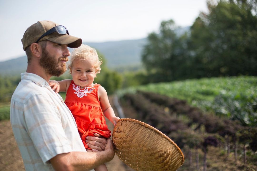 A man holds a smiling toddler in a red dress outdoors on a farm with rows of crops, mountains, and a woven basket visible.