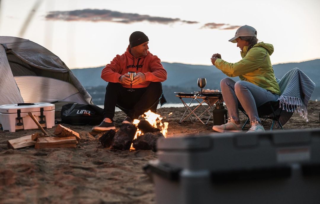 Man and woman camping on a beach at dusk, sitting near a fire with food and drinks, mountains in the background, cooler in foreground.