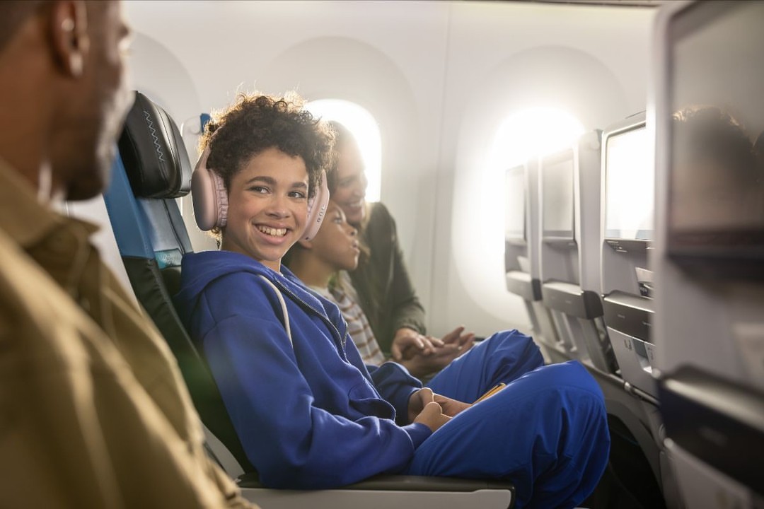 Boy in blue jumpsuit smiles on airplane wearing pink headphones, with family in background, sunlight streaming through window.