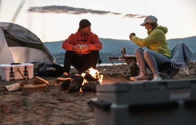 Man and woman camping by a beach campfire at sunset with a tent and portable camp chairs.