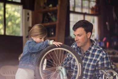 A young girl and a man working together on a bicycle wheel inside a rustic wooden garage or workshop space.