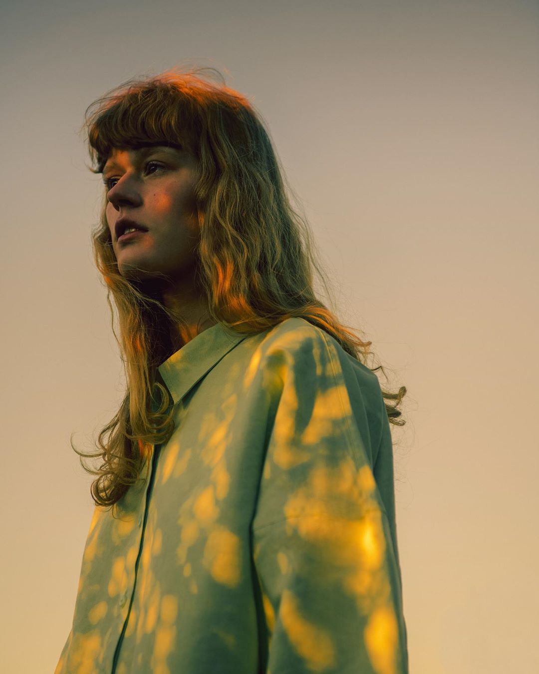Young woman with wavy blonde hair, wearing a sage green button-down shirt, is posing outdoors with a warm orange sky in the background.