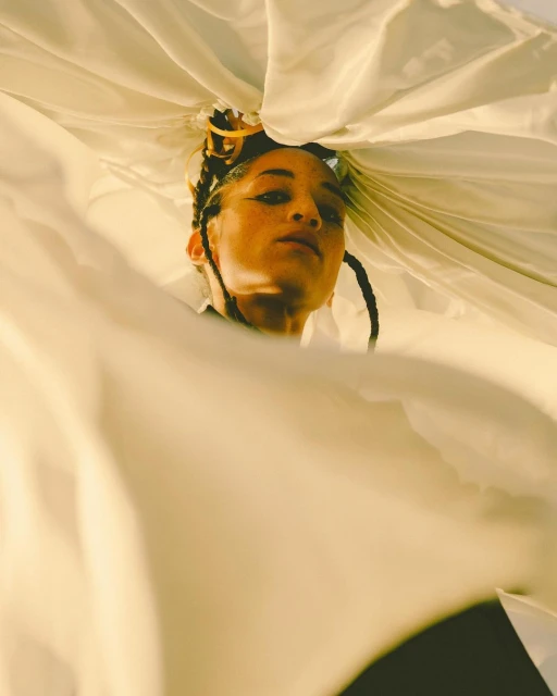 Woman with braided hair poses under a white draped fabric, creating a unique frame around her face in a low angle fashion portrait.