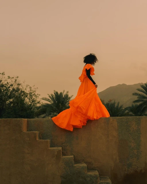 Woman in an orange gown descends stairs against a backdrop of mountains and sky at dusk, creating a vibrant, surreal landscape.