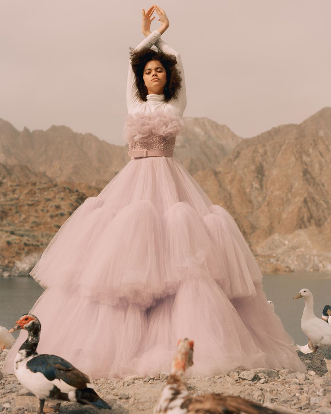 A woman in an ornate pale-purple gown poses with her arms raised in front of a landscape of mountains and ducks near a body of water.