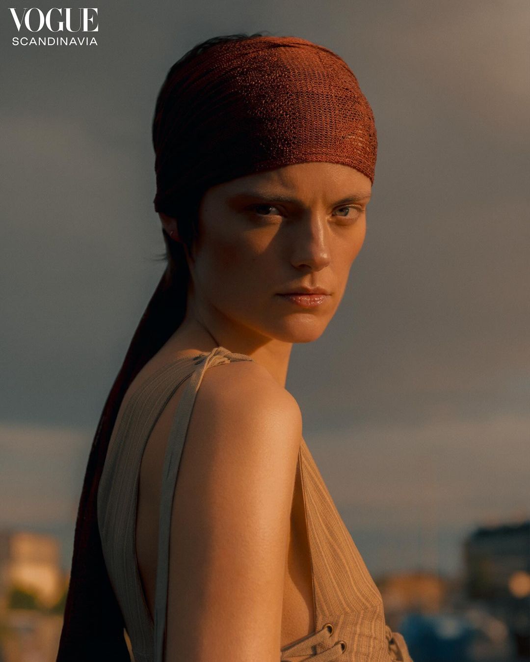 Portrait of a striking woman with a brown headscarf and pinstripe top against a moody sky. Vogue Scandinavia logo visible.