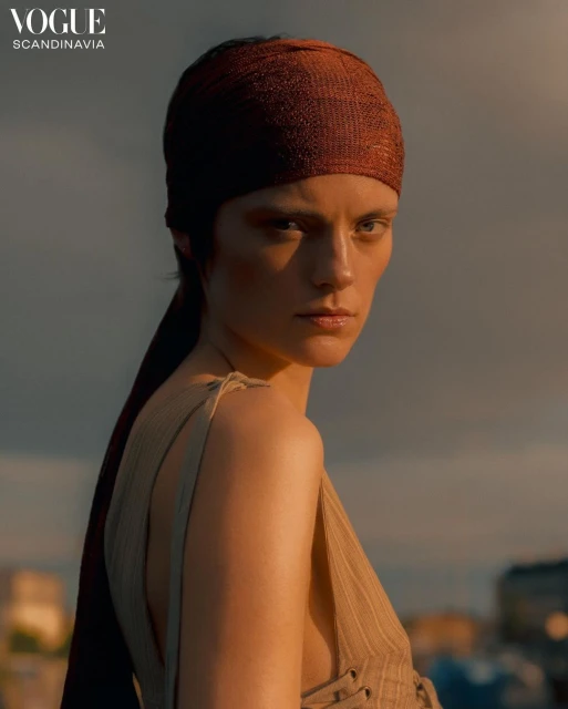 Portrait of a striking woman with a brown headscarf and pinstripe top against a moody sky. Vogue Scandinavia logo visible.