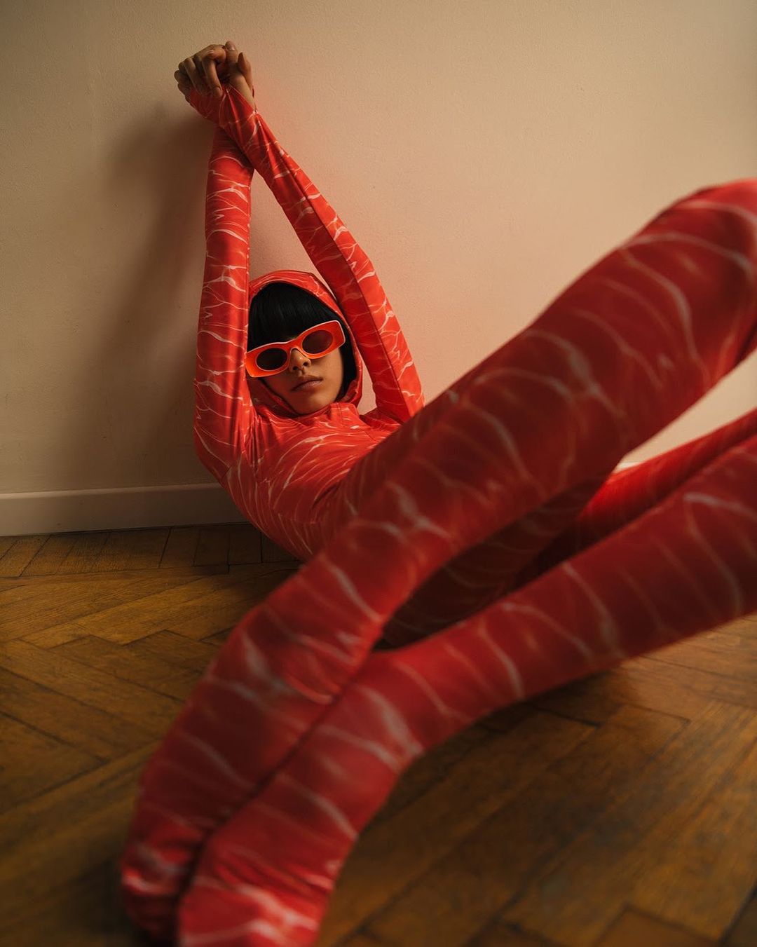 Stylish person in salmon-print jumpsuit leans against a wall indoors, accessorized with vivid orange sunglasses, on wood flooring.