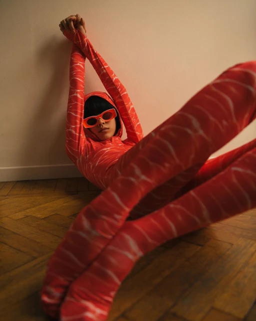 Stylish person in salmon-print jumpsuit leans against a wall indoors, accessorized with vivid orange sunglasses, on wood flooring.