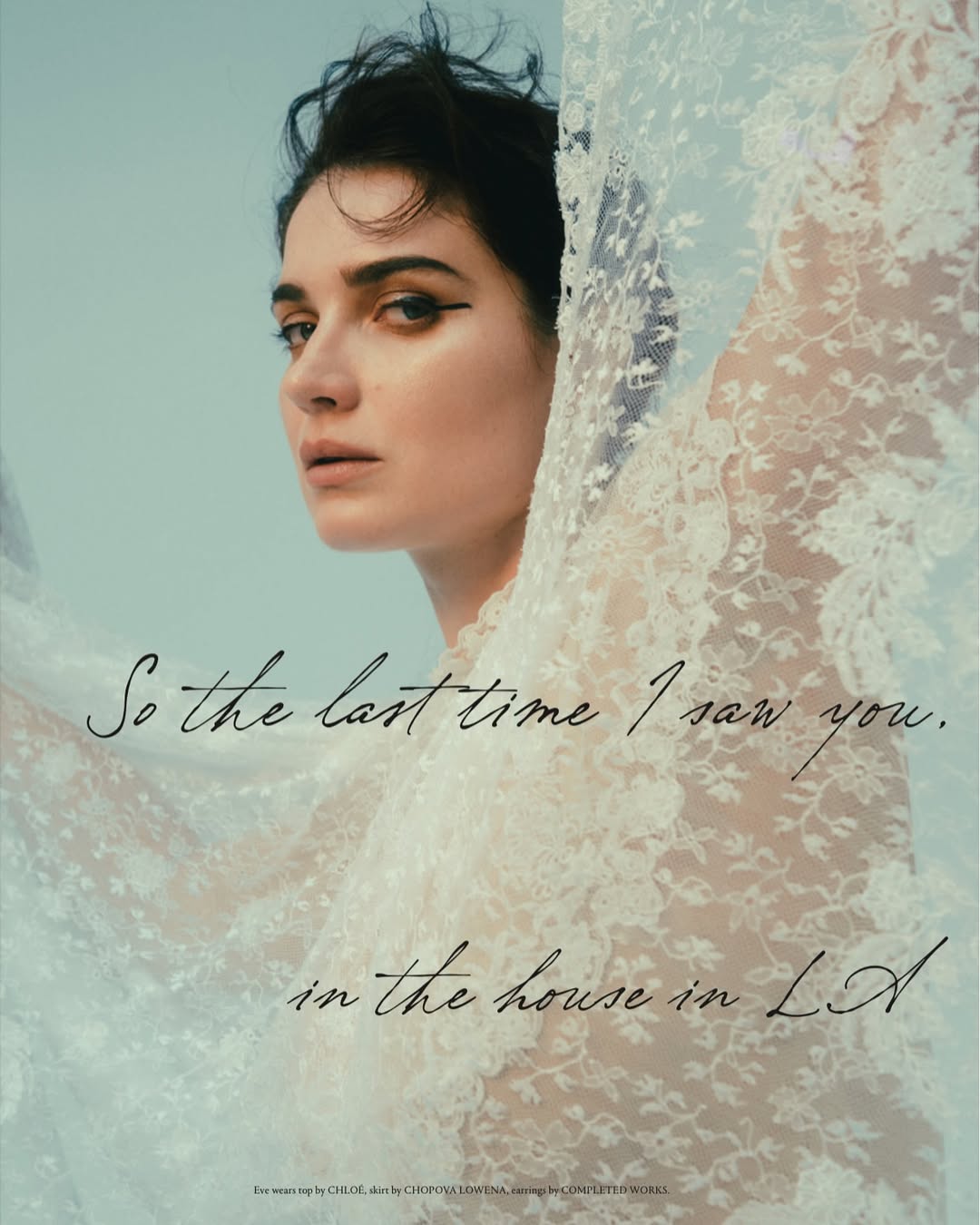 Portrait of a woman with dark hair and dramatic eyeliner posing with a lacy white fabric against a pale blue studio backdrop.