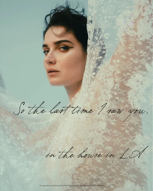 Portrait of a woman with dark hair and dramatic eyeliner posing with a lacy white fabric against a pale blue studio backdrop.