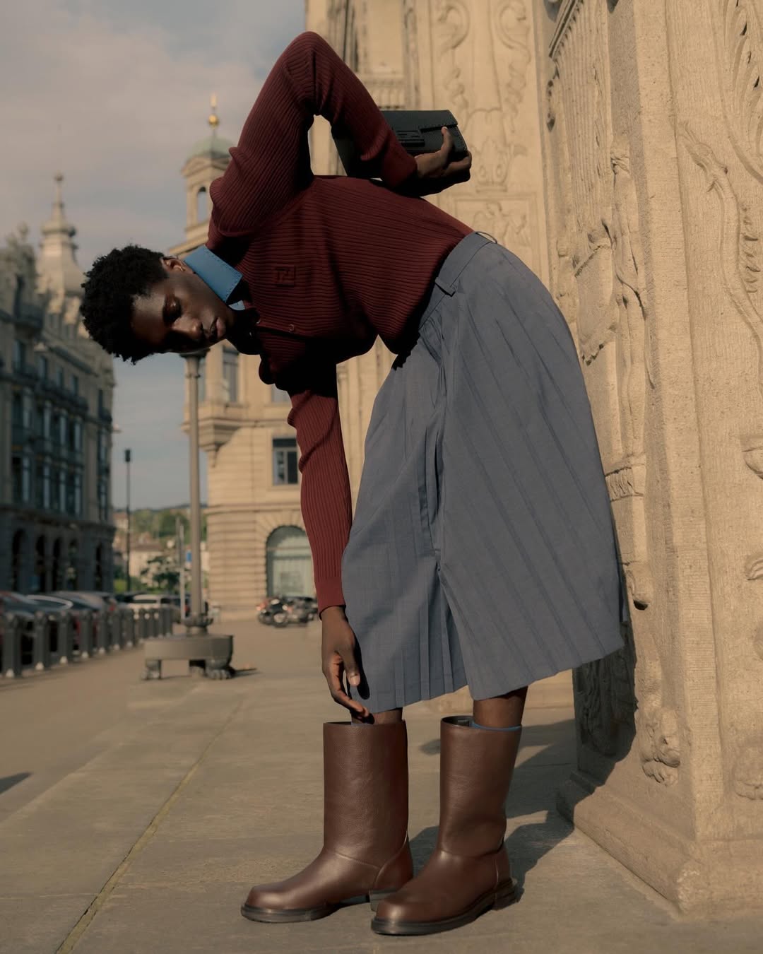 A black man in boots and a red sweater bends over in an outdoor urban setting with buildings in the background on a sunny day.