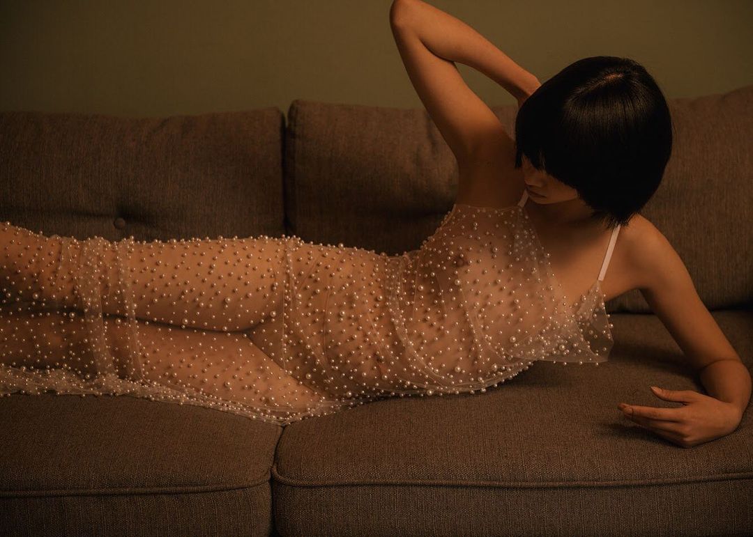An adult female with short black hair poses on a grey sofa, wearing a light dress with pearls. Indoor setting and soft, diffuse lighting.