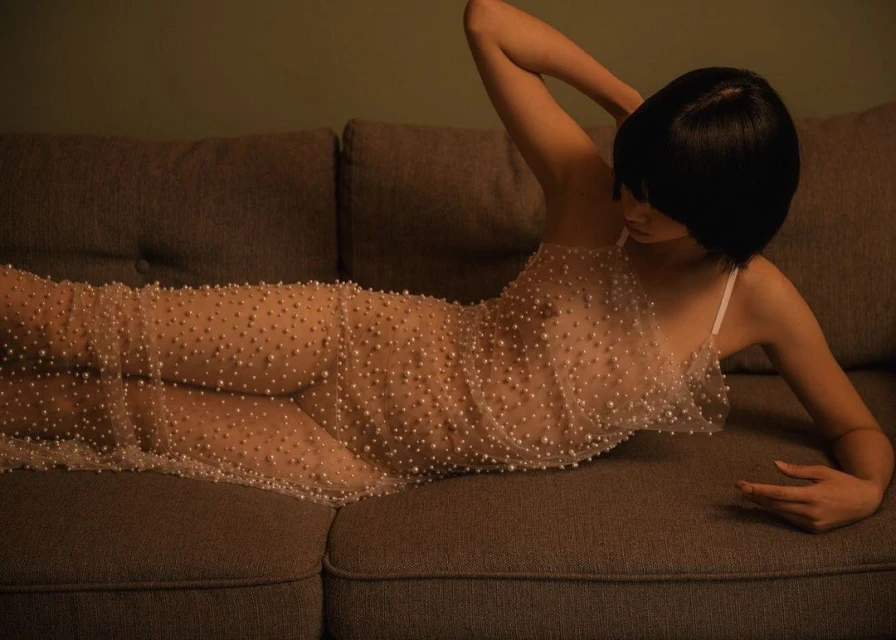 An adult female with short black hair poses on a grey sofa, wearing a light dress with pearls. Indoor setting and soft, diffuse lighting.