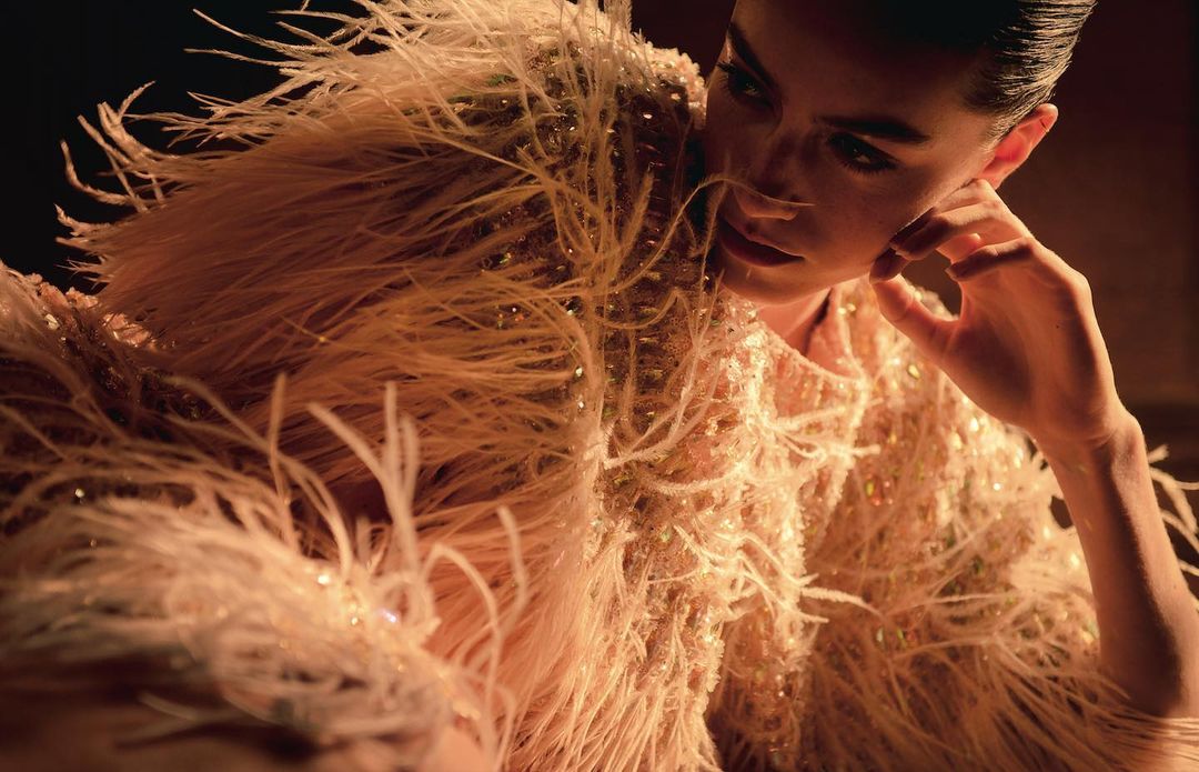 Elegant woman with hand to face, wearing feathered couture in warm, moody light, soft focus, beauty editorial. Glamour aesthetic.